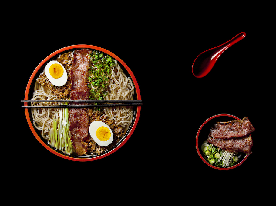 Un bol de Classic Ramen, pregătit cu grijă pentru servire, plin de tăieței de ramen și supă de carne și garnisit cu felii de bacon tras la tigaie, ceapă verde, praz și două jumătăți de ou fiert.